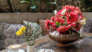 A rustic outdoor seating area with a table set for coffee and pastries, surrounded by greenery and a stone wall.