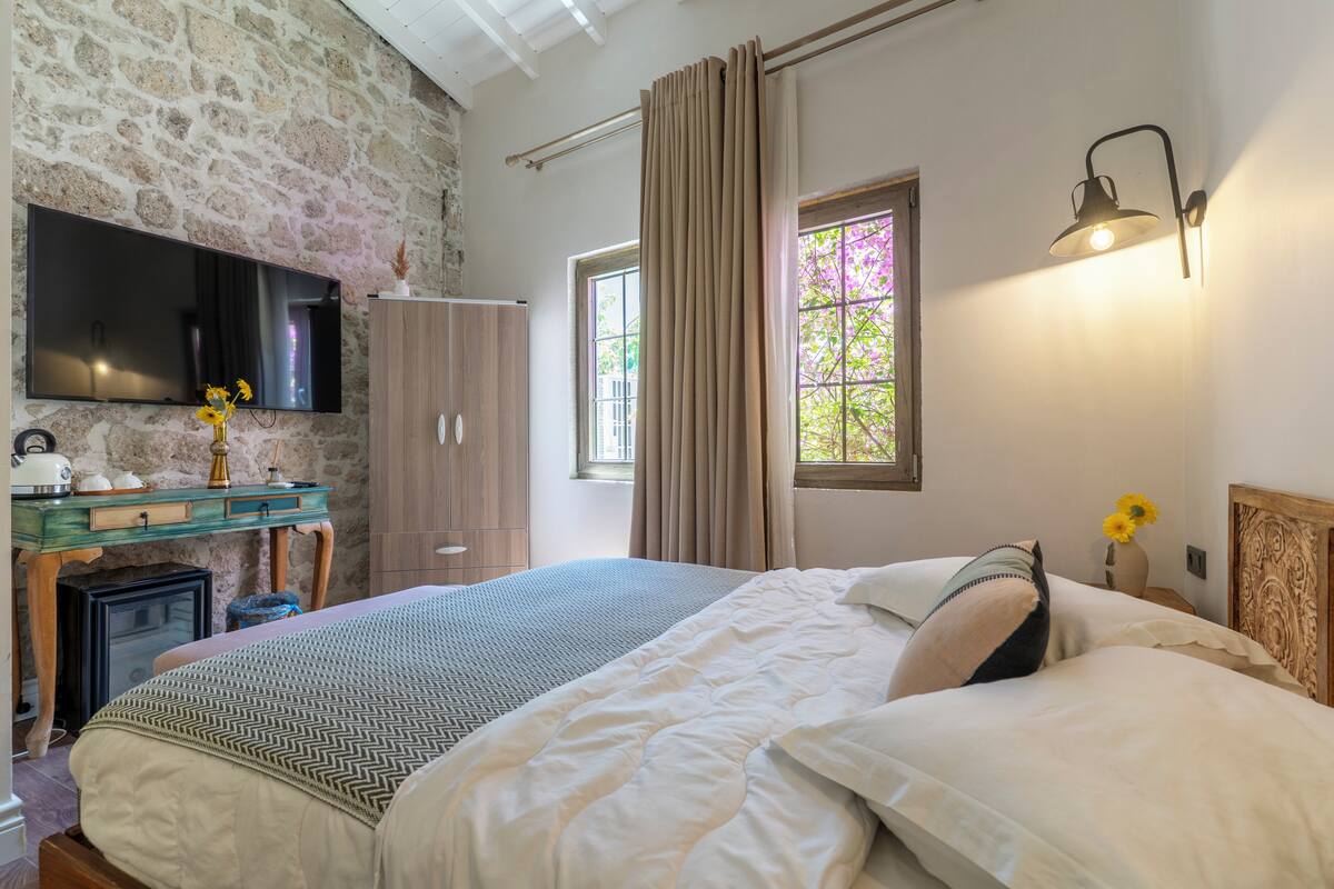 Un dormitorio con una cama grande, un aparador, un televisor montado en una pared de piedra y una ventana con cortinas.