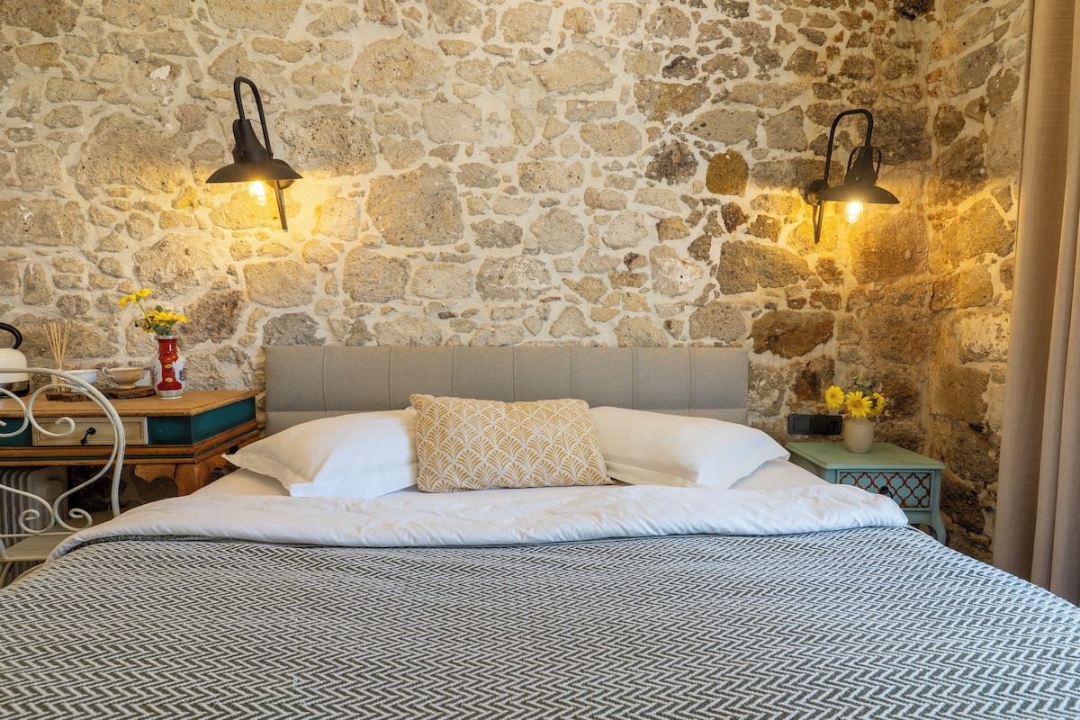Un dormitorio con pared de piedra, una cama con ropa de cama estampada, dos lámparas fijadas a la pared y una mesita de noche con un jarrón de flores.