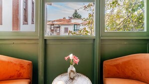 A room with two orange chairs, a small table with a vase of flowers, a cup of coffee, and a view of buildings through a window. - Mediha Hanım Konağı Alaçatı (Cesme)