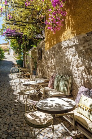 Una calle empedrada con mesas y sillas para exteriores, junto a una pared de piedra cubierta de plantas trepadoras.