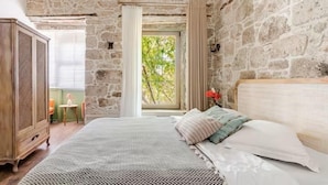 A bedroom with a stone wall, a bed with patterned bedding, a wooden wardrobe, and a window with curtains. - Mediha Hanım Konağı Alaçatı (Cesme)