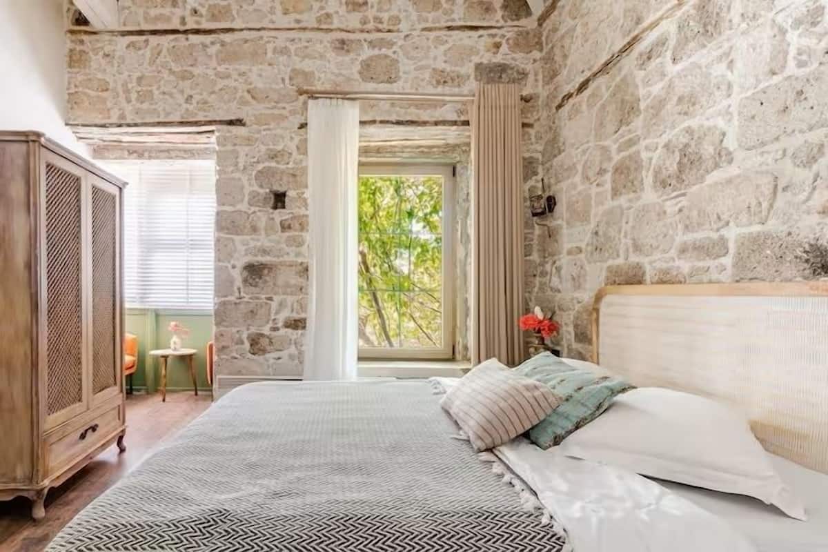 Un dormitorio con pared de piedra, cama con ropa de cama estampada, armario de madera y ventana con cortinas.