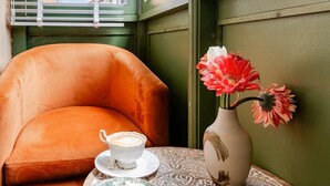 A cozy seating area with a cushioned chair, a small round table with a patterned tablecloth, a white cup, a plate with pastries, and a vase with flowers. - Mediha Hanım Konağı Alaçatı (Cesme)