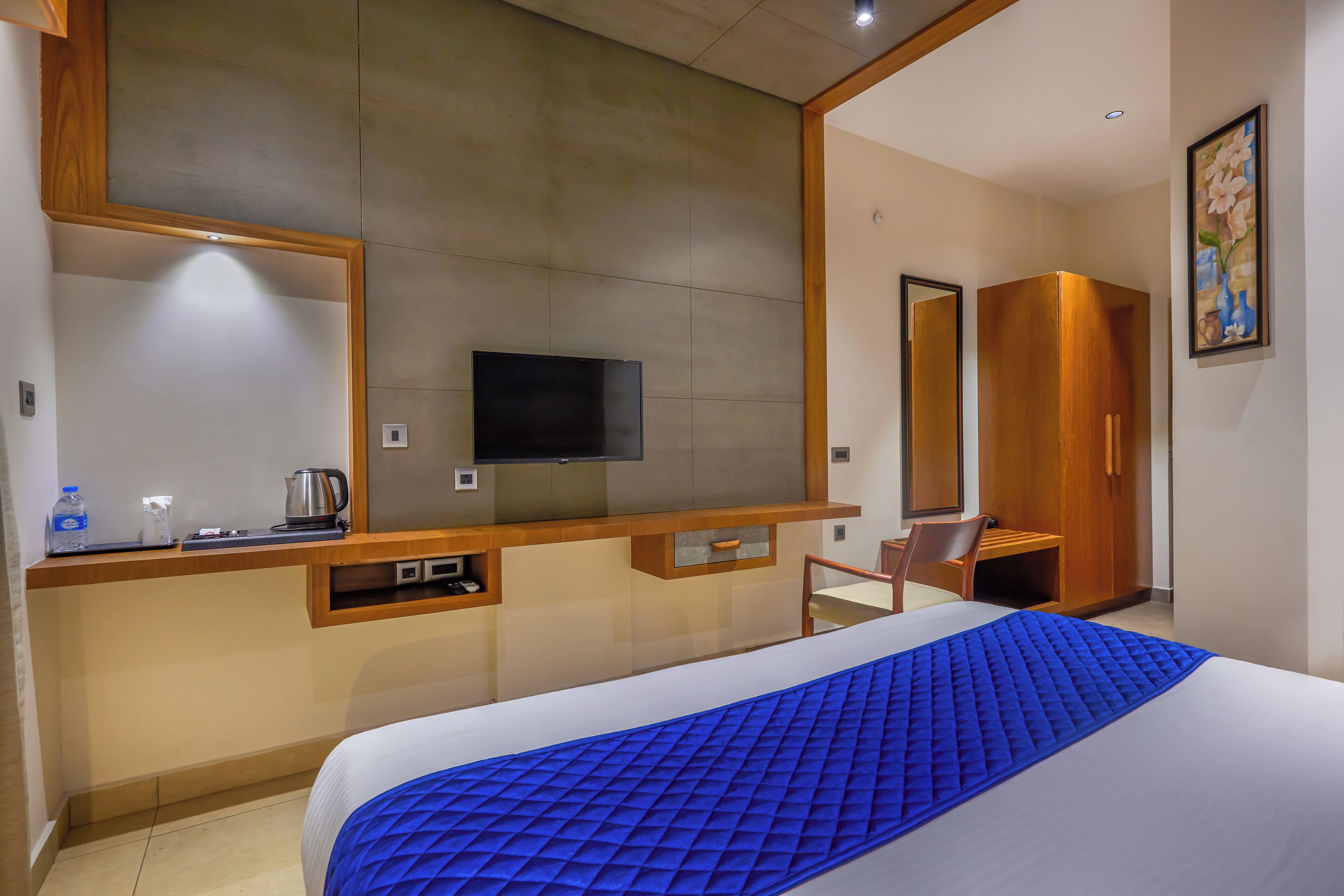 A hotel room with a bed, a TV mounted on the wall, a wooden desk, a chair, and a wardrobe.