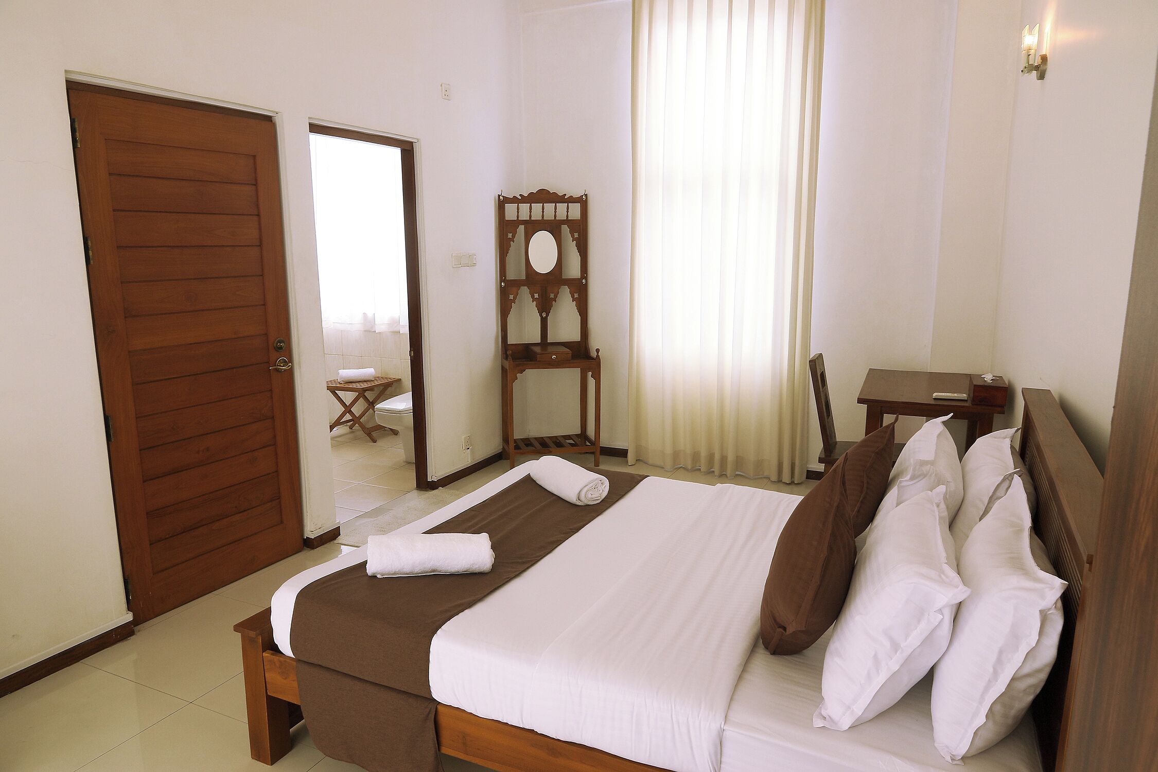 Une chambre d’hôtel avec un lit en bois, une chaise en bois, une petite table et un miroir au mur.
