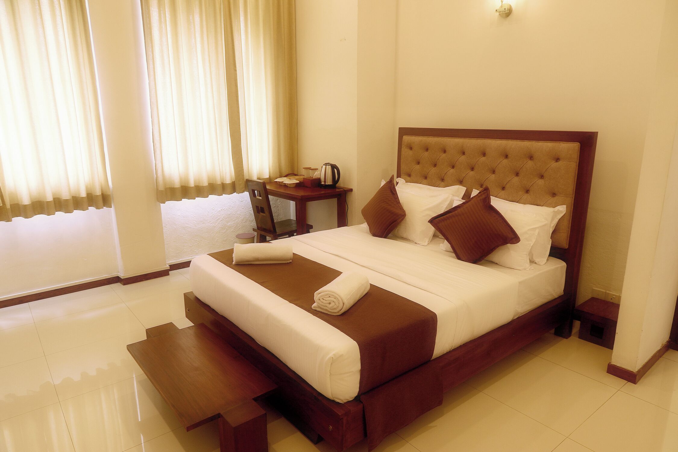 A hotel room with a large bed, a bedside table, a chair, and a small table with a kettle.