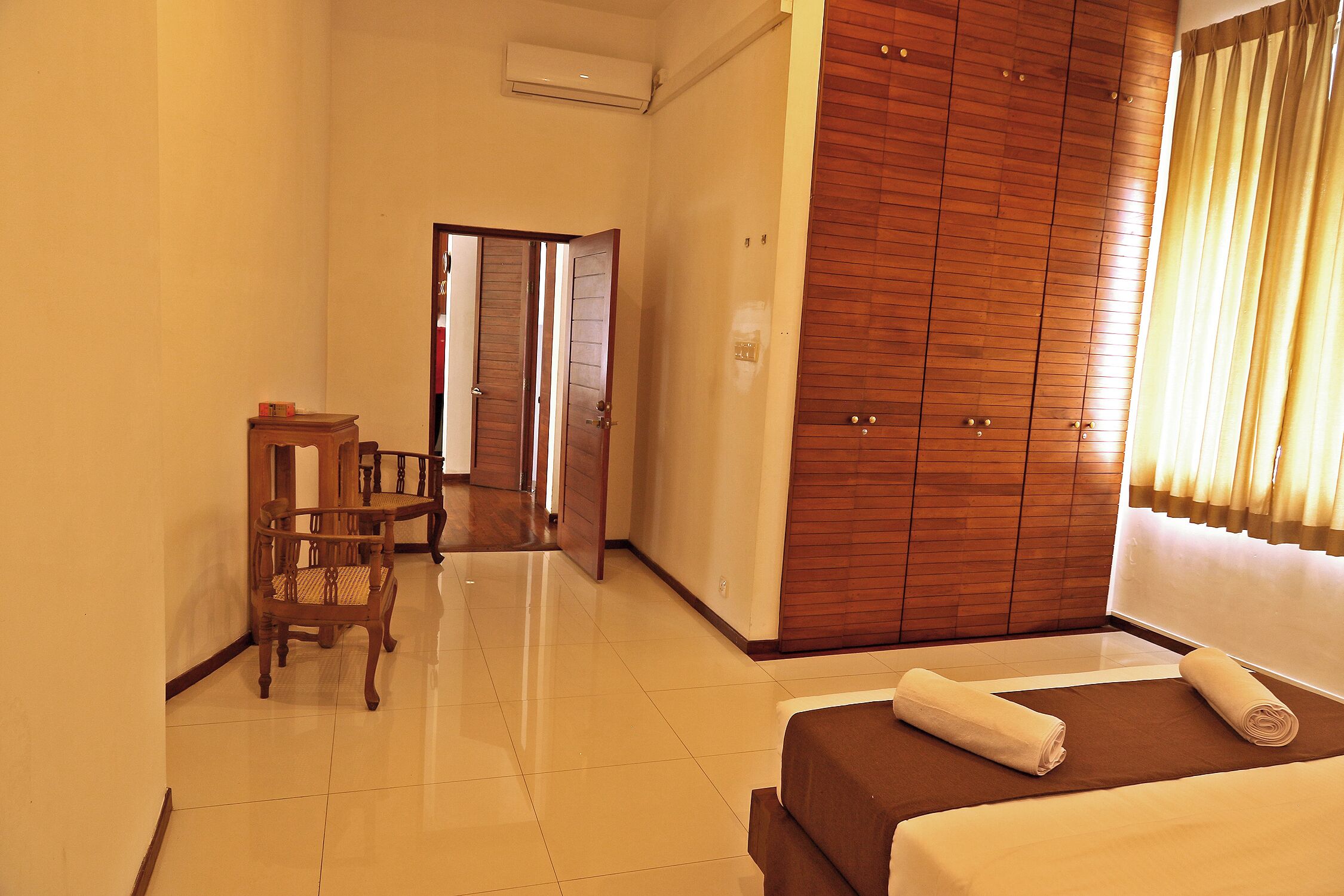 A hotel room with a bed, a wooden wardrobe, a small table with chairs, and a door leading to another room.