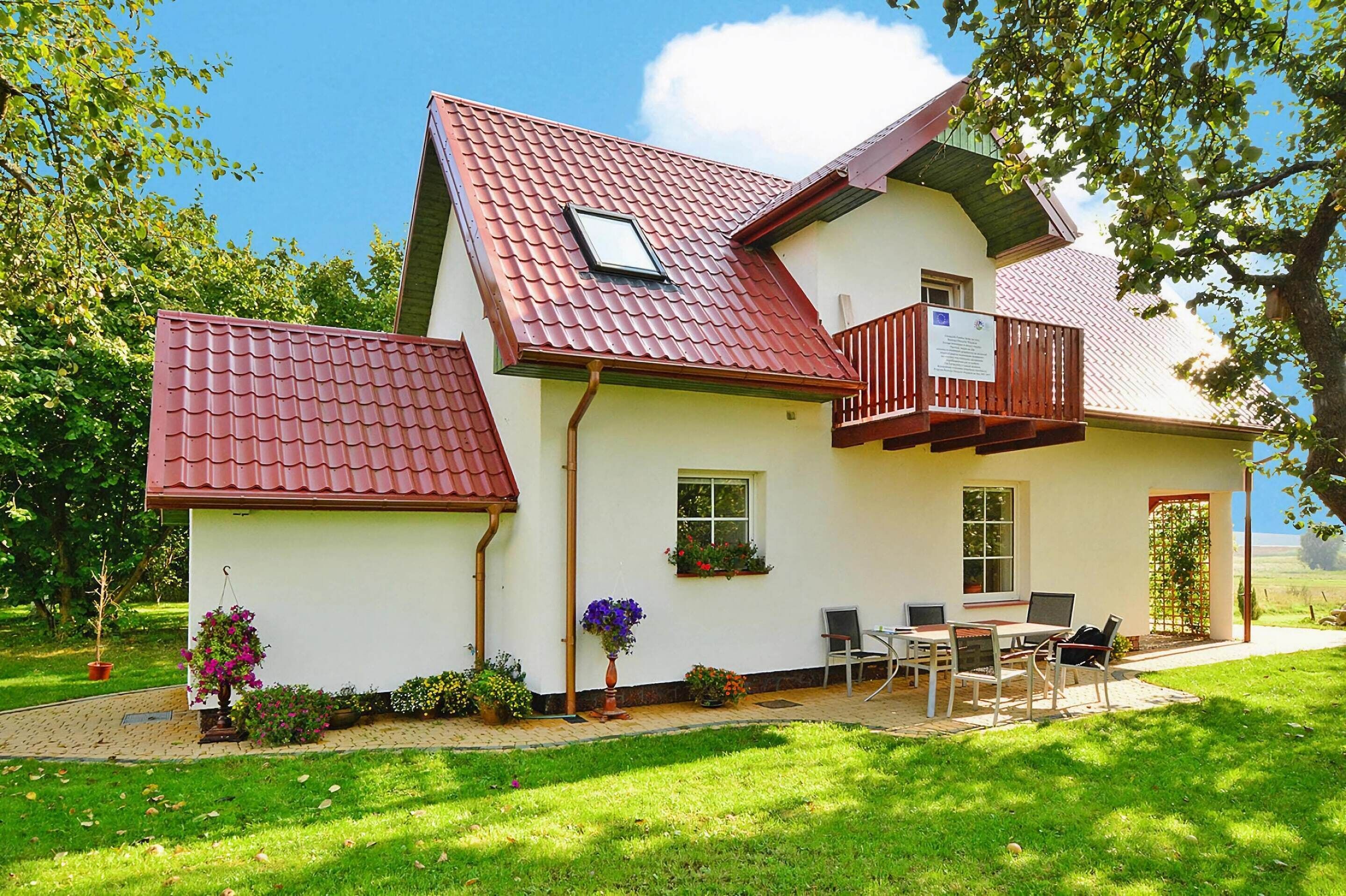 A house with a red-tiled roof, a balcony, a patio with a table and chairs, and a garden with potted plants.
