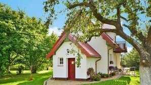A small white house with a red door and a balcony, surrounded by lush greenery and a paved path. - Large Holiday Home With a Beautiful Garden (Slawno)