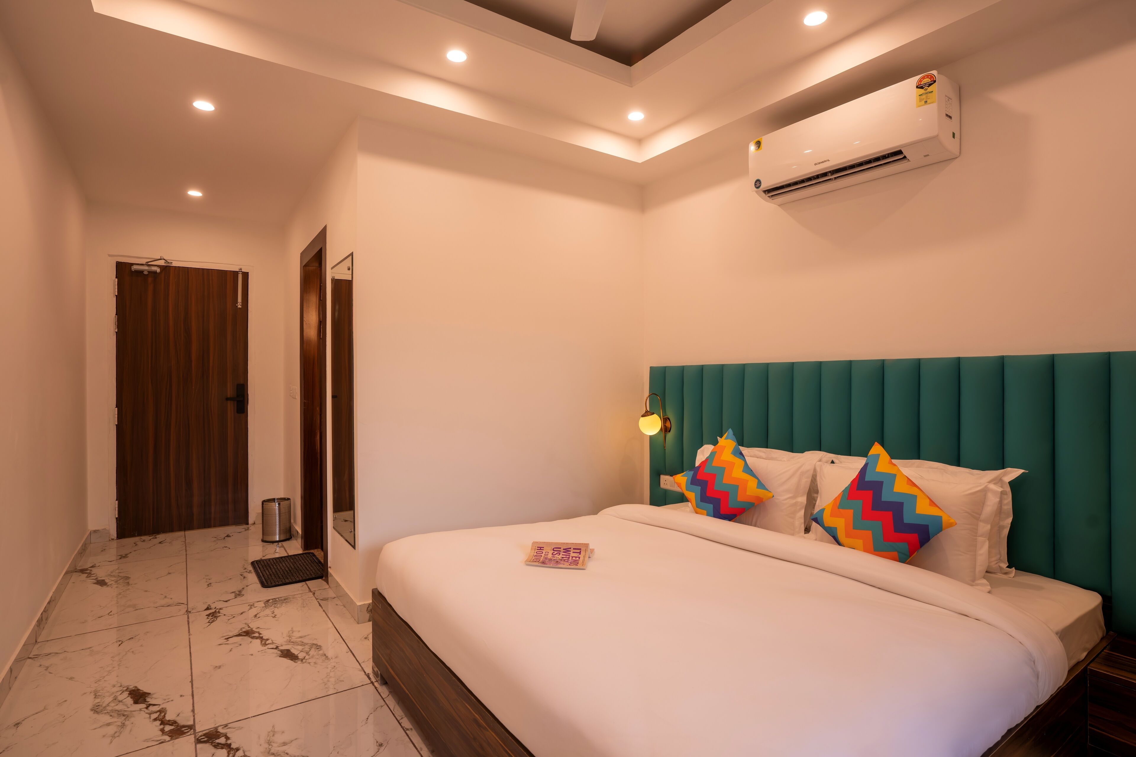 A hotel room with a bed, a wooden door, a wall-mounted air conditioner, and a ceiling with recessed lighting.