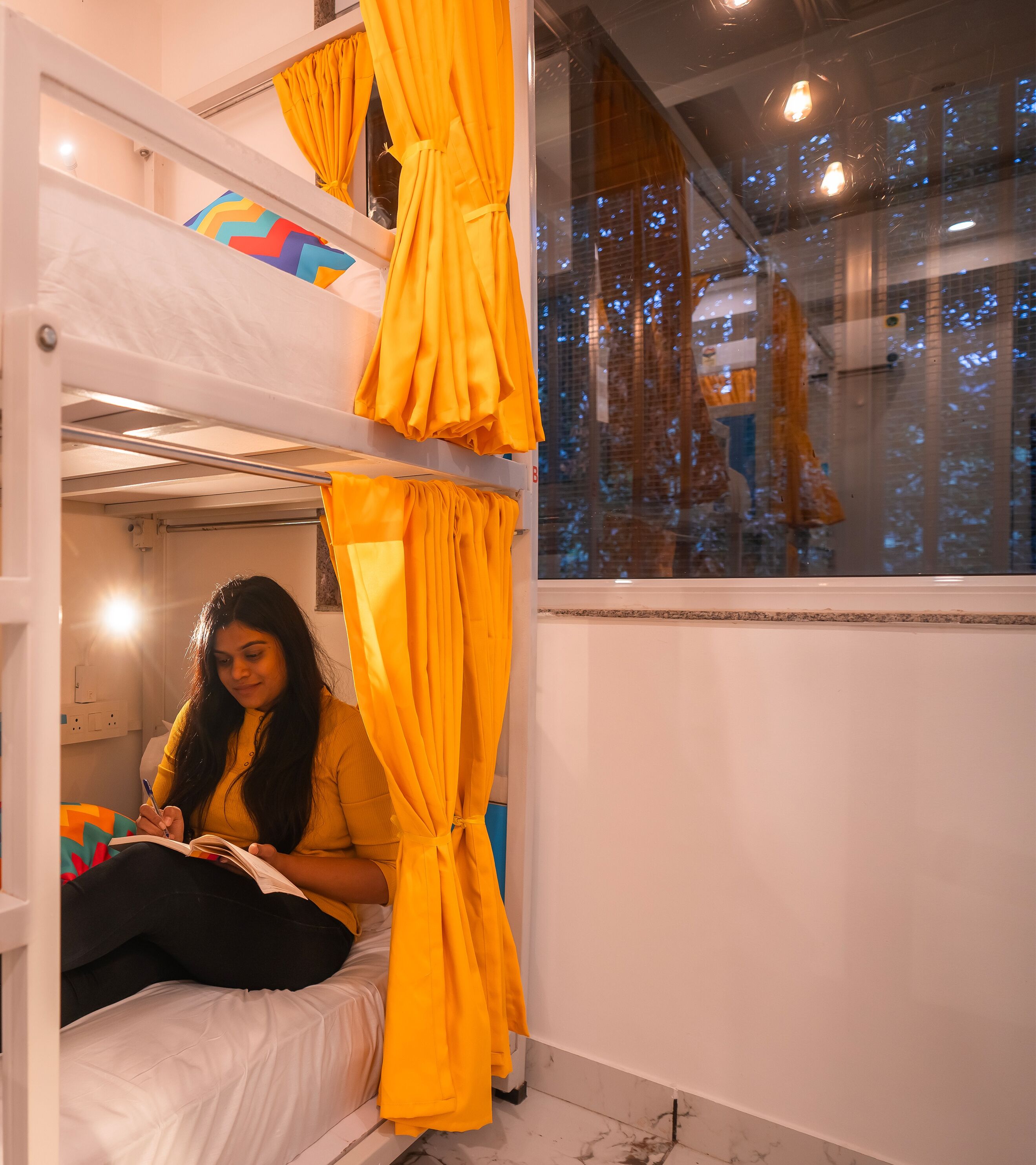 Une chambre avec un lit superposé, une fenêtre, des rideaux jaunes et une personne en train d’écrire.