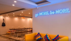 Et moderne hotelværelse med et neonskilt der siger "go MORE. be MORE." og en levende gul sofa med farverige puder.