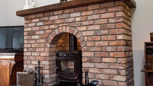 A brick fireplace with a wooden mantel, a television, and a wicker basket. - Nelly Daly's Barn - Sleeps 6 - Brand New - Parking (Newry)