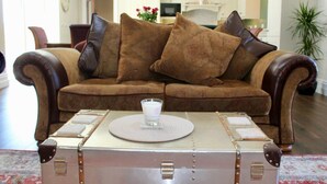 A brown leather sofa with matching cushions, a vintage suitcase on a round table, and a kitchen in the background. - Nelly Daly's Barn - Sleeps 6 - Brand New - Parking (Newry)