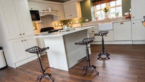 A kitchen with white cabinets, a marble countertop, and a wooden floor. There are three bar stools with metal baskets. - Nelly Daly's Barn - Sleeps 6 - Brand New - Parking (Newry)