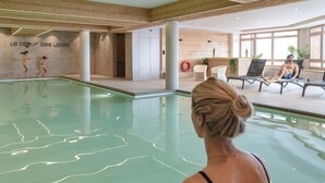 Ein Hallenbad mit freier Sicht zum Beckenrand, eine Holzvertäfelung mit einem Schild und eine Sitzecke mit Liegestühlen und einer entspannten Person.