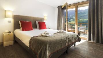 Quarto de hotel com uma cama grande, duas luminárias de cabeceira, uma mesinha de madeira e uma varanda com vista para o campo.