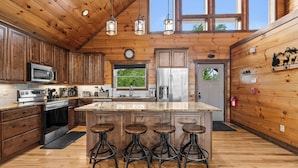 A spacious kitchen with wooden cabinets, a large island with stools, stainless steel appliances, and a window overlooking greenery. - Private Indoor Pool, Hot Tub, Free Attractions, Pool Table, Fire Pit (Gatlinburg)