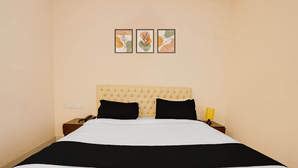 A neatly made bed with a tufted headboard, a bedside table with a lamp, and three framed artworks on the wall. - Classic HBR Layout Formerly Leela Park (Bengaluru)