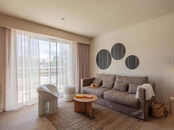 Un salón moderno con un sofá gris, sillones blancos y una mesa de centro. Hay tres adornos redondos en la pared y un gran ventanal con cortinas de gasa.