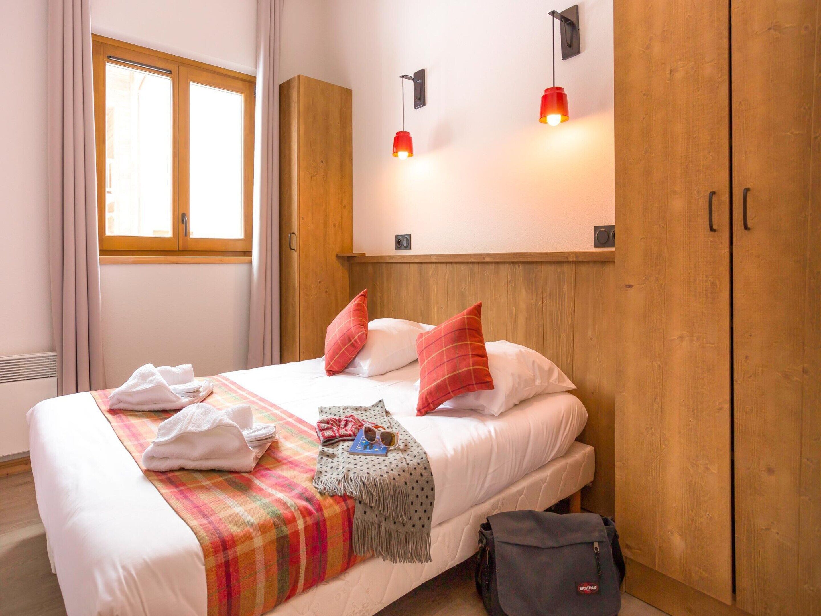 A hotel room with a bed, a wooden wardrobe, a window, and two wall-mounted lights.