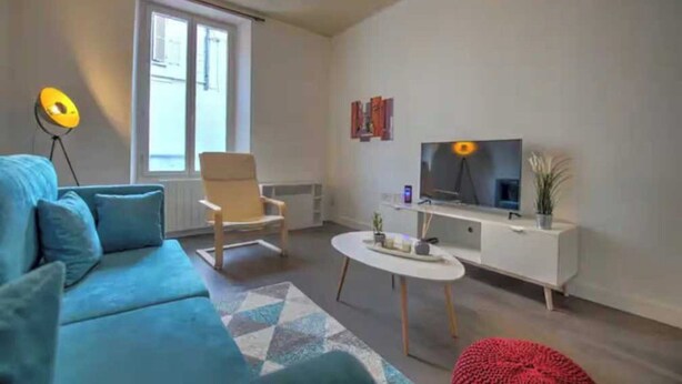 Uma sala de estar moderna com um sofá azul, uma mesa de centro branca, um banquinho vermelho, uma televisão sobre um móvel branco e um abajur.