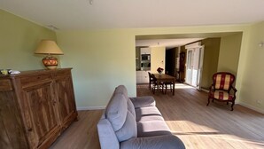 Uma sala de estar com sofá cinza, armário de madeira e uma luminária. Dá para ver uma área de jantar com mesa e cadeiras ao fundo.