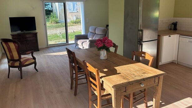 Uma área de refeições com mesa e cadeiras de madeira, uma televisão e uma cozinha equipada com frigorífico e máquina de lavar louça.