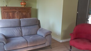Uma sala de estar com um sofá cinzento, uma poltrona vermelha, um armário de madeira e uma máquina de lavar roupa ao fundo.