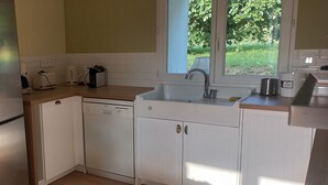 Cozinha compacta com armários brancos, geladeira de aço inoxidável, lava-louças e janela com vista para o verde lá fora.