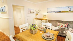 Un coin repas avec une table en bois, des chaises blanches et un centre de table floral. On y trouve également un canapé avec un coussin à motifs floraux et une lampe posée sur une petite table en bois.