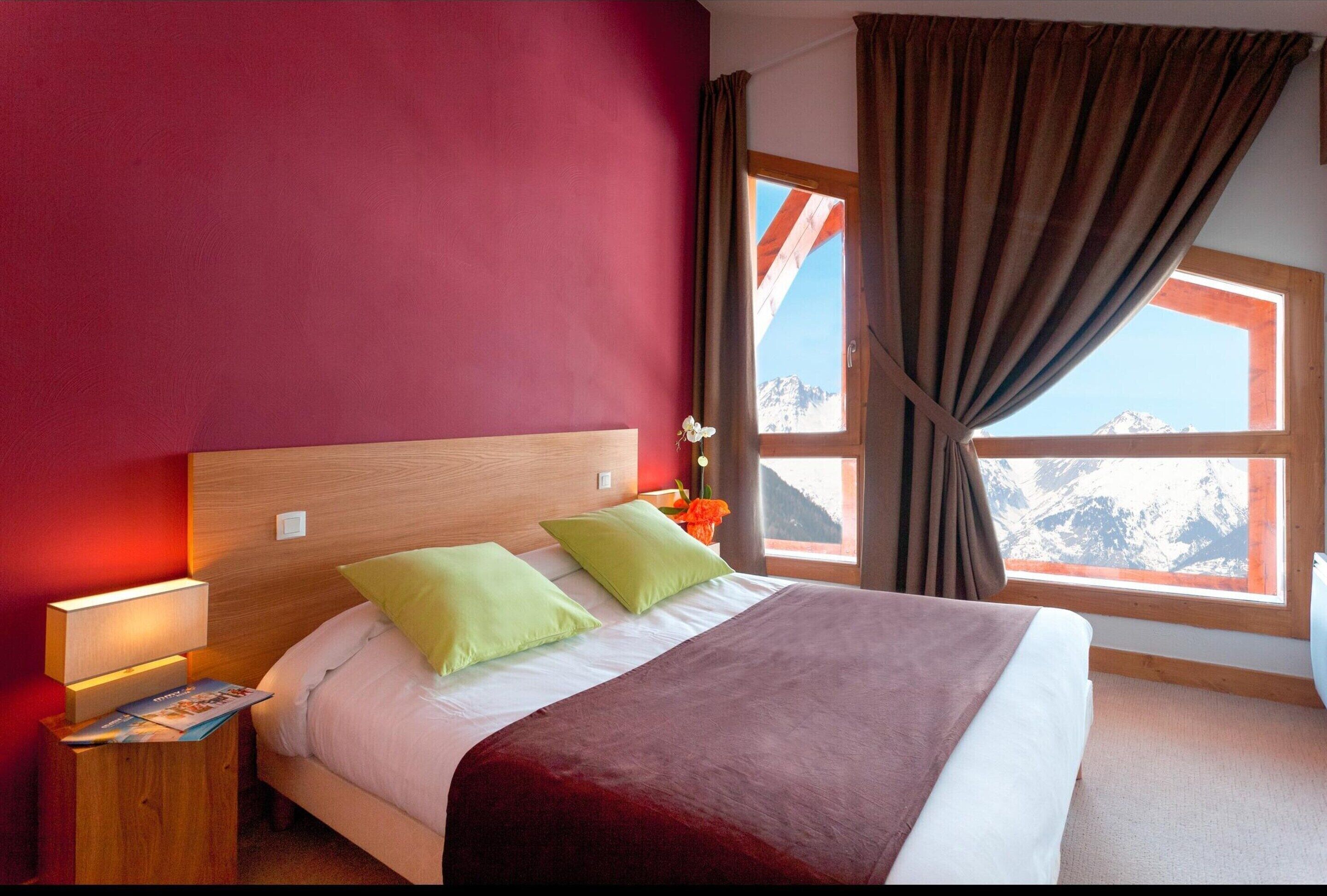 Une chambre d’hôtel avec un grand lit, des murs rouges, une tête de lit en bois et une fenêtre donnant sur la montagne.