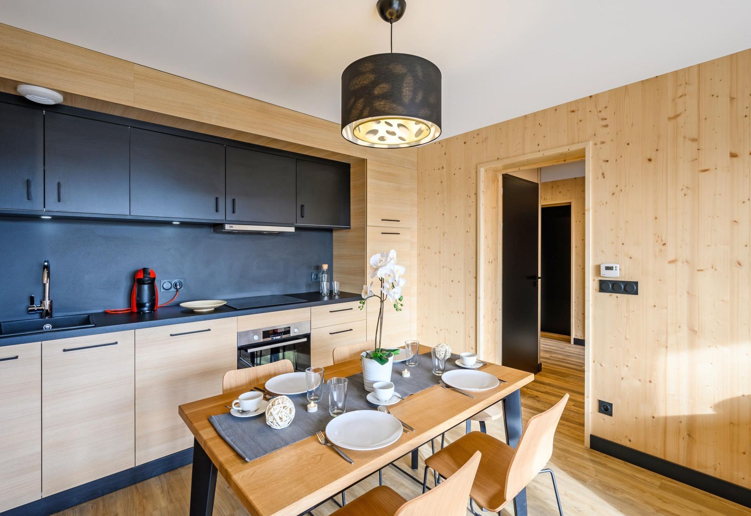 A modern kitchen with dark cabinets, a wooden dining table set for four, and a pendant light above.