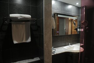 Un baño moderno con una pared de azulejos oscuros, una toalla blanca sobre un perchero metálico, un espejo grande y un lavabo con encimera de mármol.