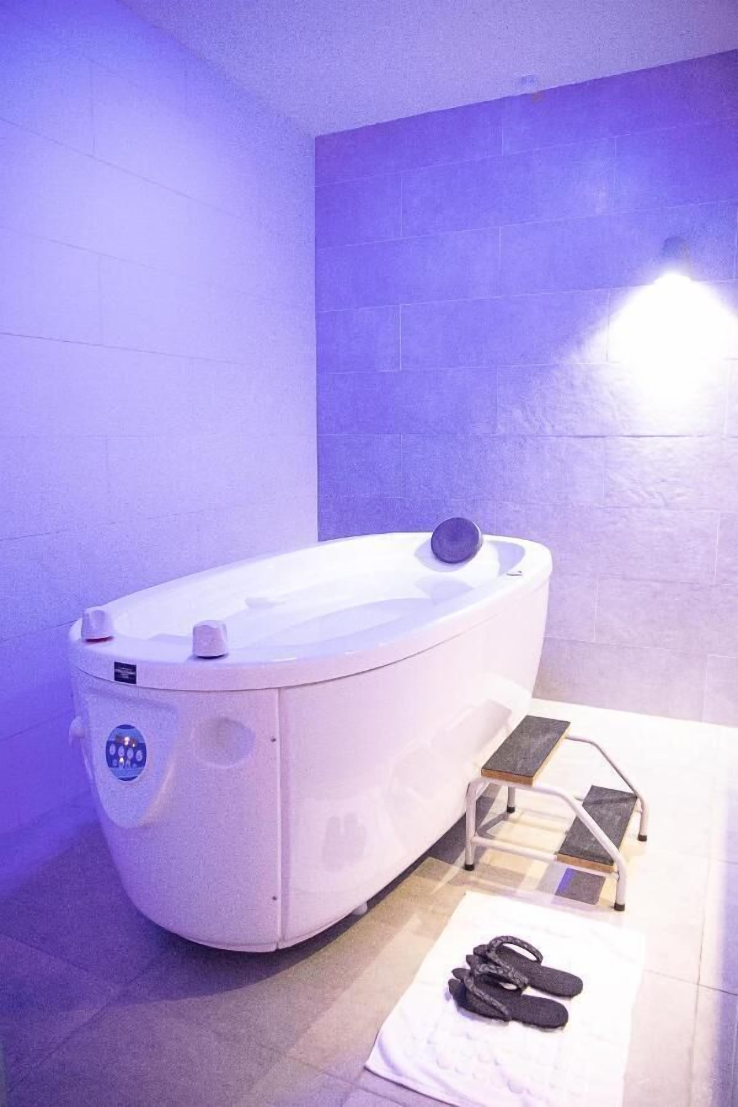 Ein Spa-Raum mit einer weißen Whirlwanne, einem Handtuchhalter und einem Paar Hausschuhen auf dem Boden.
