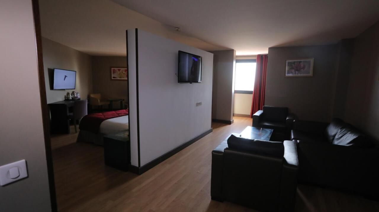 A hotel room with a bed, a TV mounted on the wall, a desk with a chair, and a seating area with a sofa and armchairs.