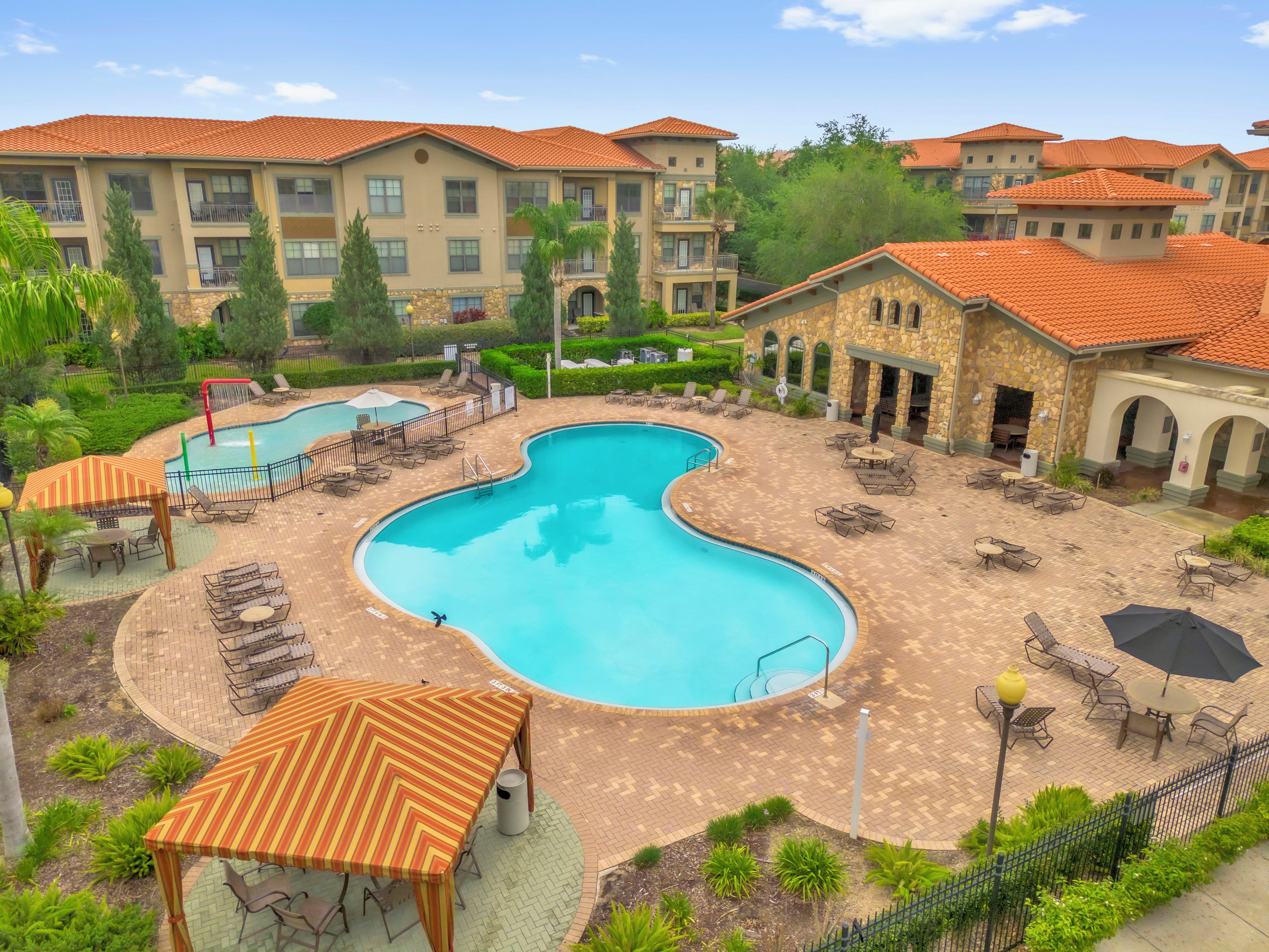 A swimming pool with lounge chairs and umbrellas, surrounded by a brick patio and a stone building with a tiled roof.
