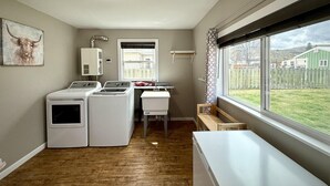 Ein Waschraum mit zwei Frontlader-Waschmaschinen, einem Waschbecken und einem Fenster mit Blick auf ein grünes Haus und einen Zaun.