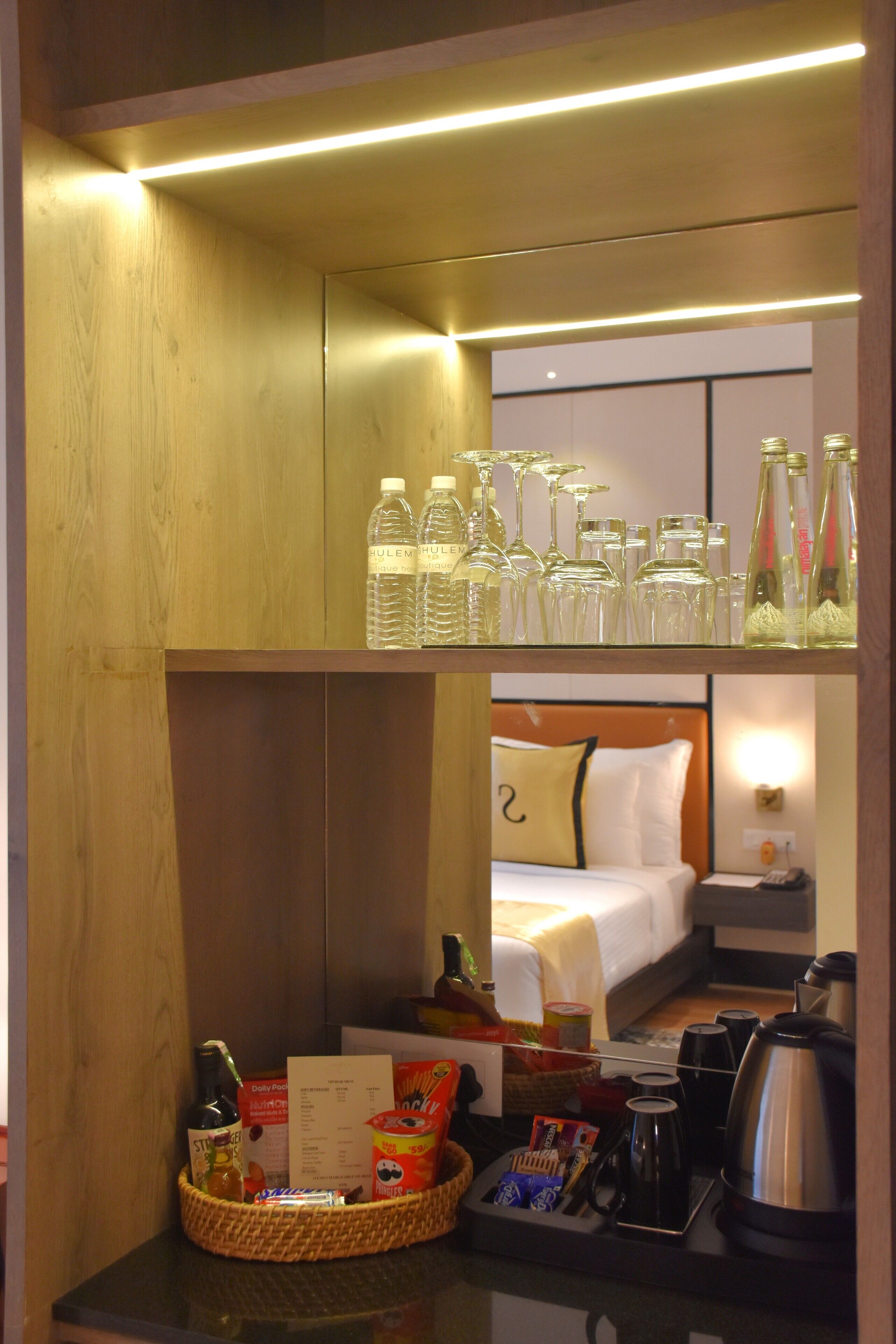 A hotel room with a wooden wall, a bed with a white and yellow pillow, a nightstand with a lamp, and a table with a basket containing snacks and a bottle.