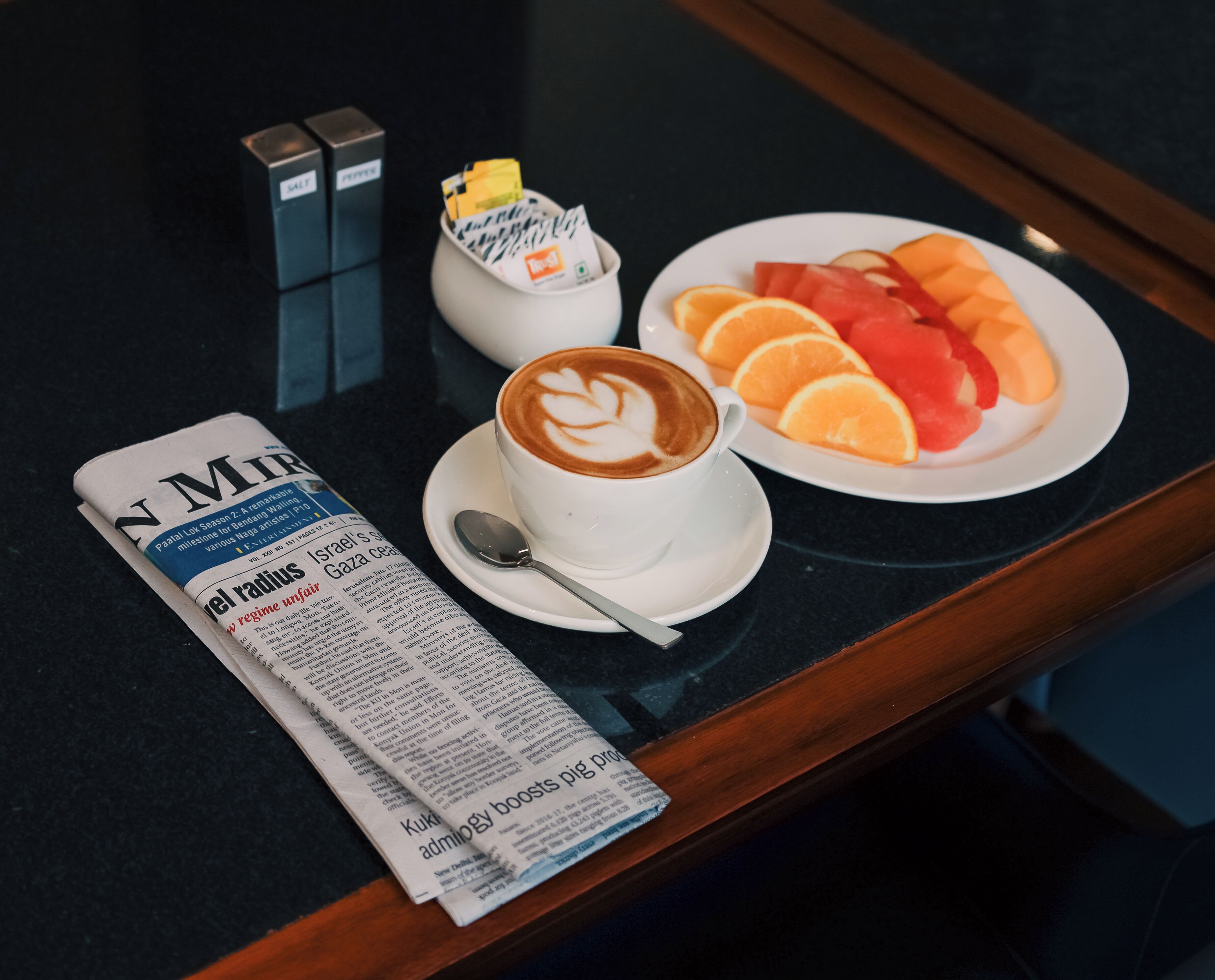 Une table avec un journal, une tasse de café décorée d’un motif en latte art, une assiette de fruits et un petit récipient contenant un sachet jaune.