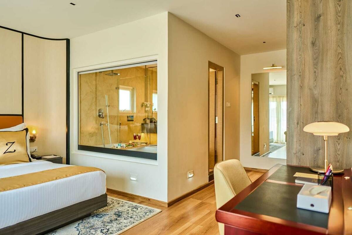 A hotel room with a large bed, a desk, a chair, a mirror, and a view of the bathroom through a glass window.