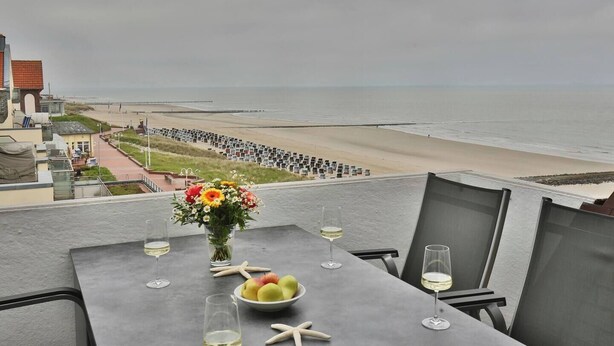 A table with a vase of flowers, a bowl of fruit, and wine glasses on a balcony overlooking a beach with a sea wall of photographs. - Beach Villa Marina 26 Sea View (Wangerooge)