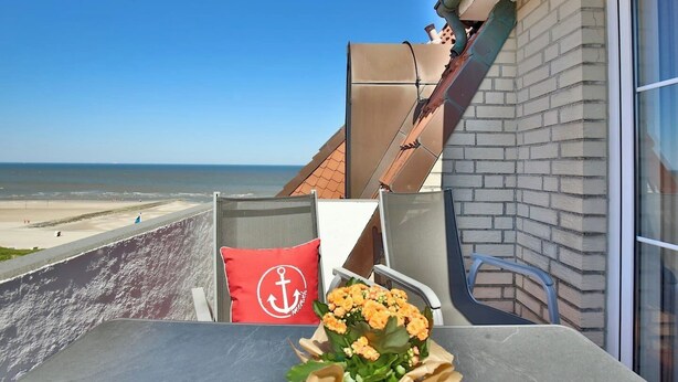 Un balcon avec vue sur la plage, une table agrémentée d’un arrangement floral et un coussin rouge orné d’un motif d’ancre.