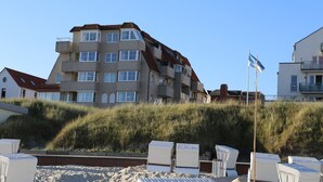 Un immeuble à plusieurs étages, doté d’un toit en pente, entouré de dunes de sable et de chaises de plage.