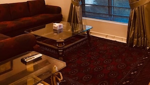Una habitación de hotel con una alfombra de color rojo oscuro con diseño, un sofá marrón oscuro, una mesa de centro de vidrio y una ventana con cortinas.