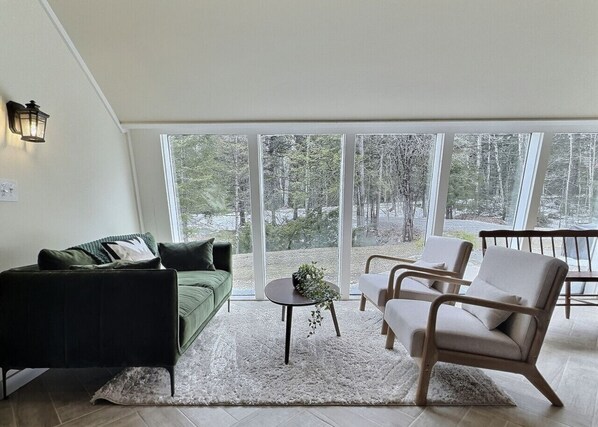 A modern living room with a green sofa, a round coffee table, and two armchairs. Large windows offer a view of trees outside. - Holiday Haus- AC, game room, near golf (Killington)
