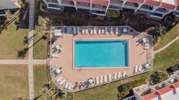 An aerial view of a rectangular swimming pool surrounded by lounge chairs and sunbeds, with a small deck area and a pathway leading to a building.