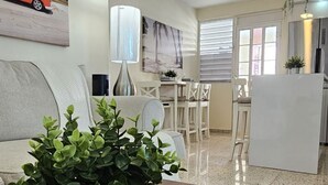Una sala de estar con sofá, una mesa de centro con una planta, un cuadro en la pared y una cocina al fondo.