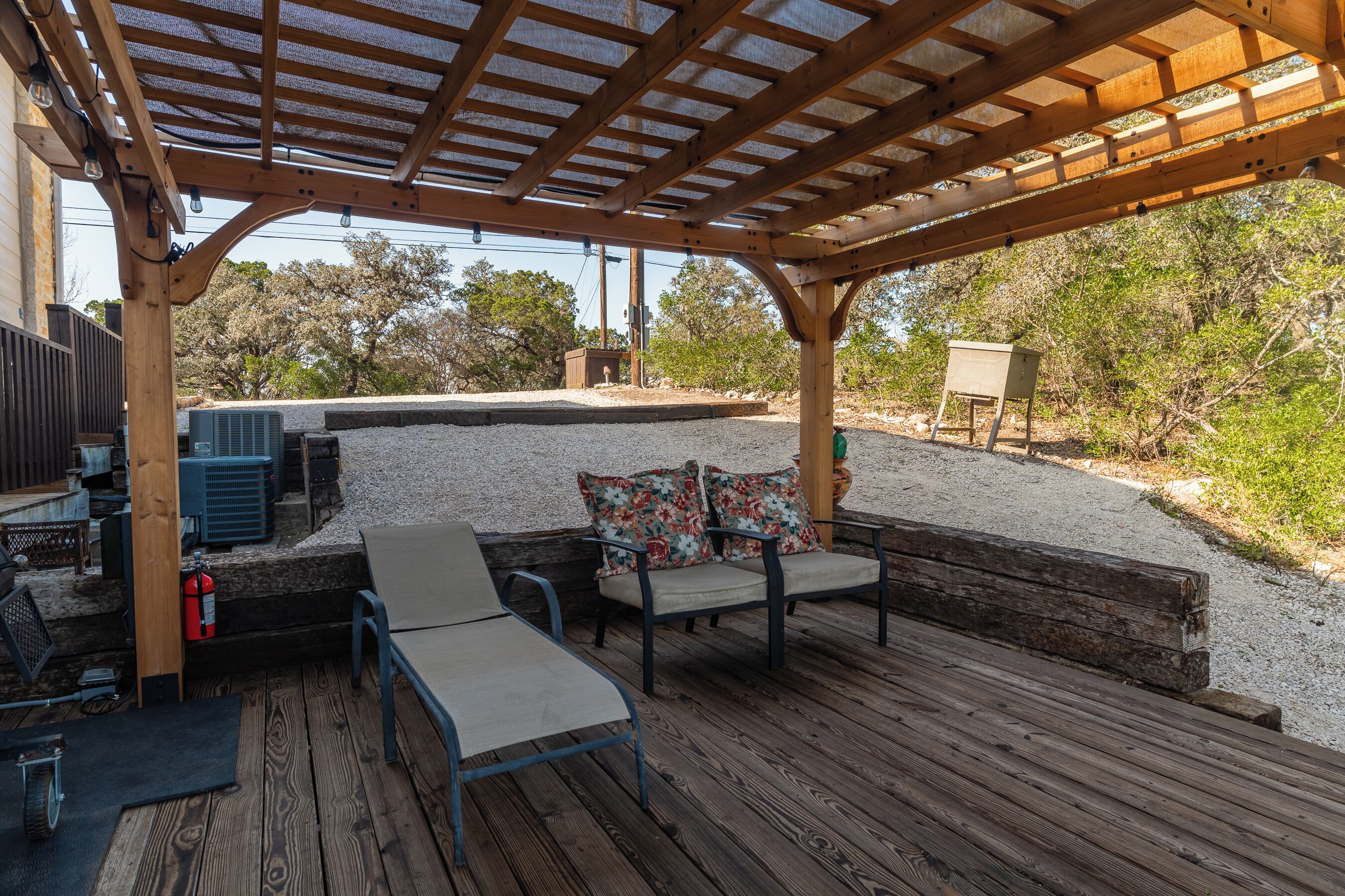 An outdoor patio with wooden flooring, a pergola with string lights, a fire extinguisher, and a seating area with cushions.